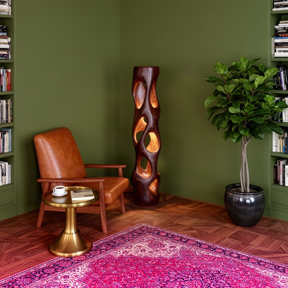 Ambera wood floor lamp glowing warmly through carved openings in an olive green reading corner with leather chair